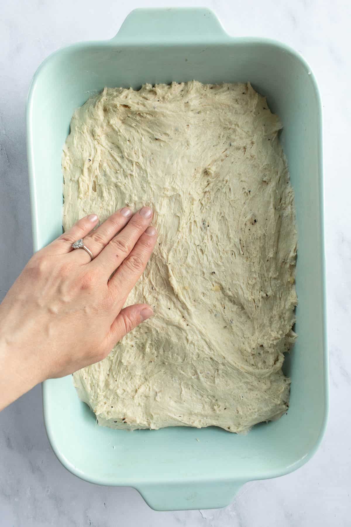 A hand pressing white bread dough into a blue baking dish.