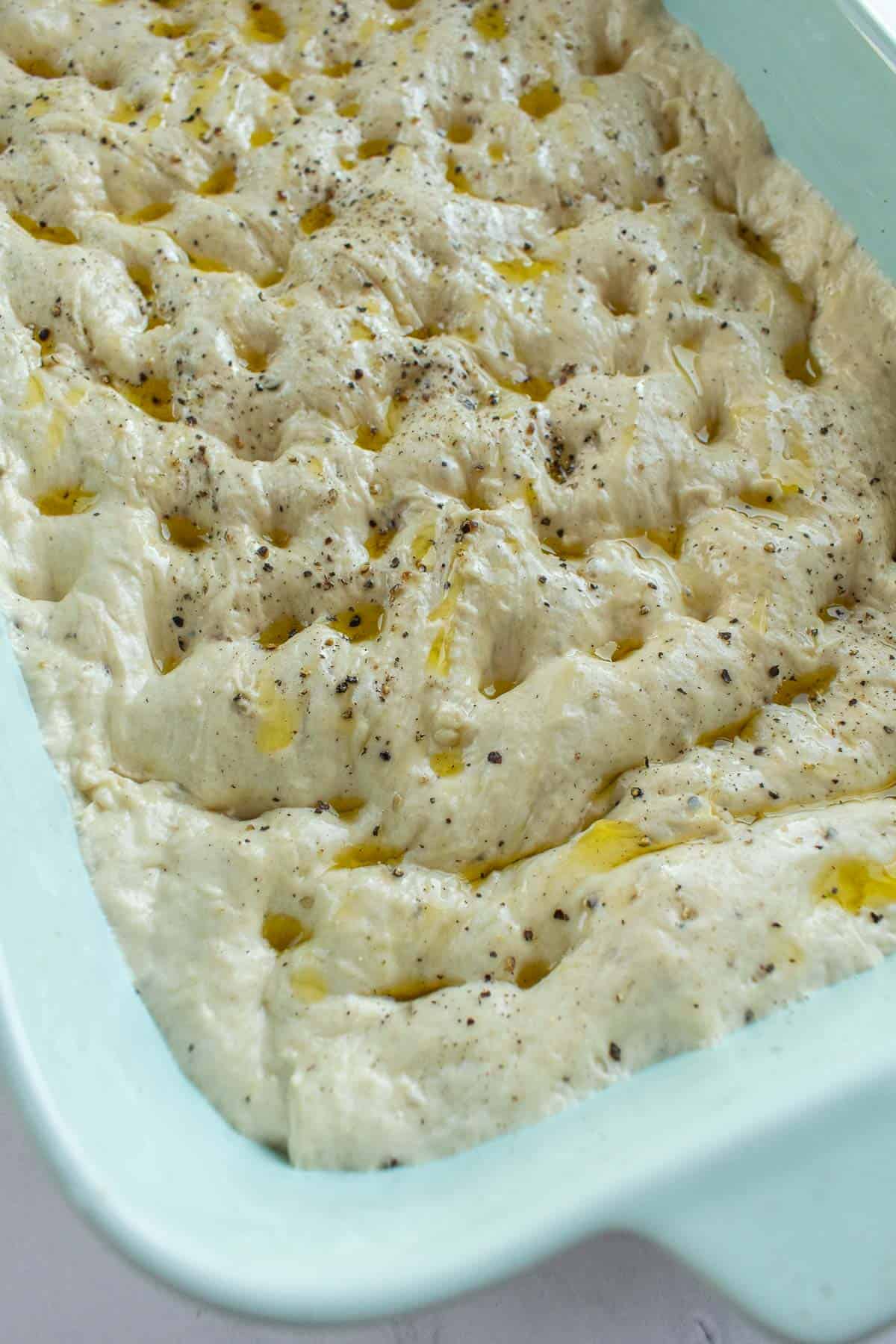 A blue baking dish filled with dimpled focaccia dough topped with black pepper and olive oil.