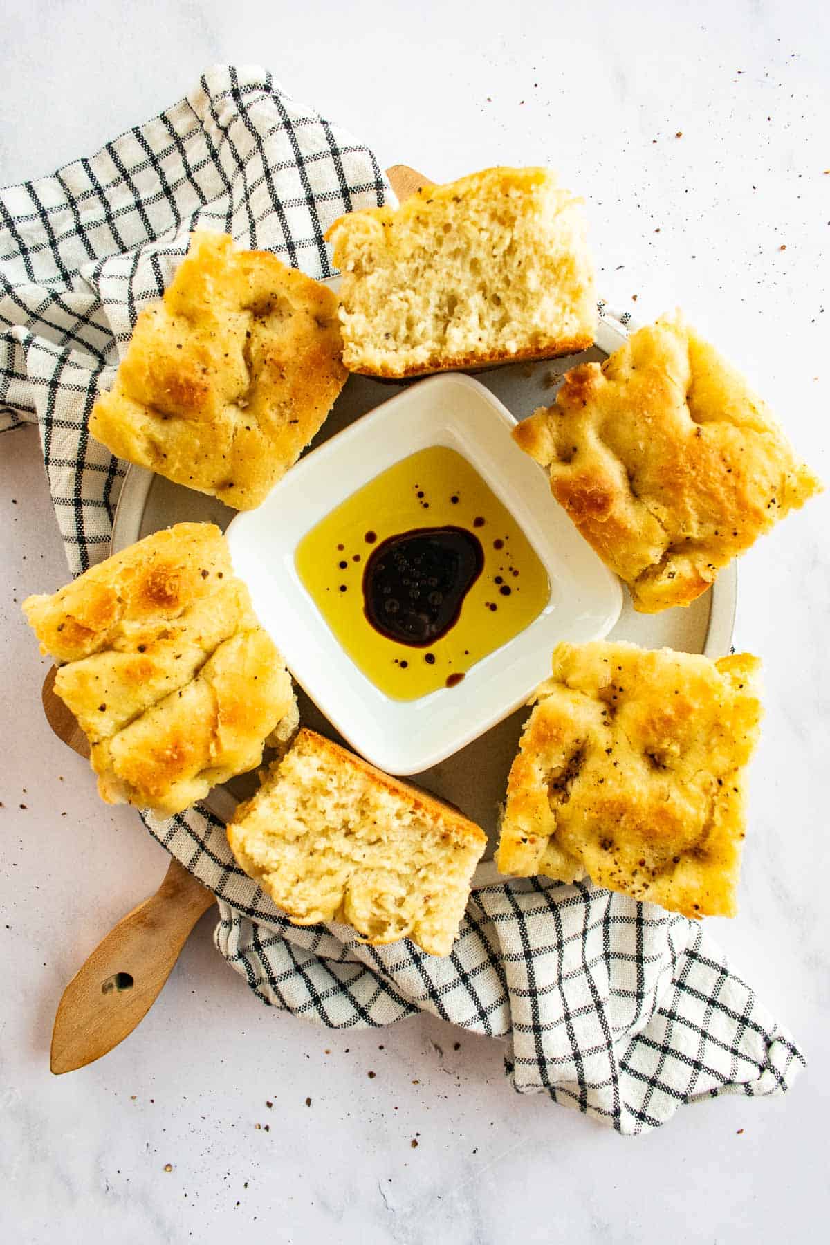 Six large squares of focaccia bread on a wooden board surround a square bowl of olive oil and vinegar.