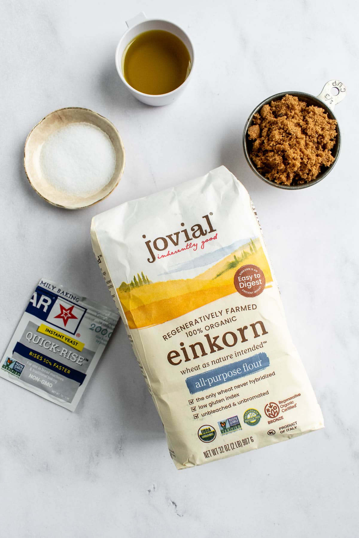 A bag of einkorn flour next to dishes of brown sugar, salt, and olive oil and a yeast packet on a light surface.