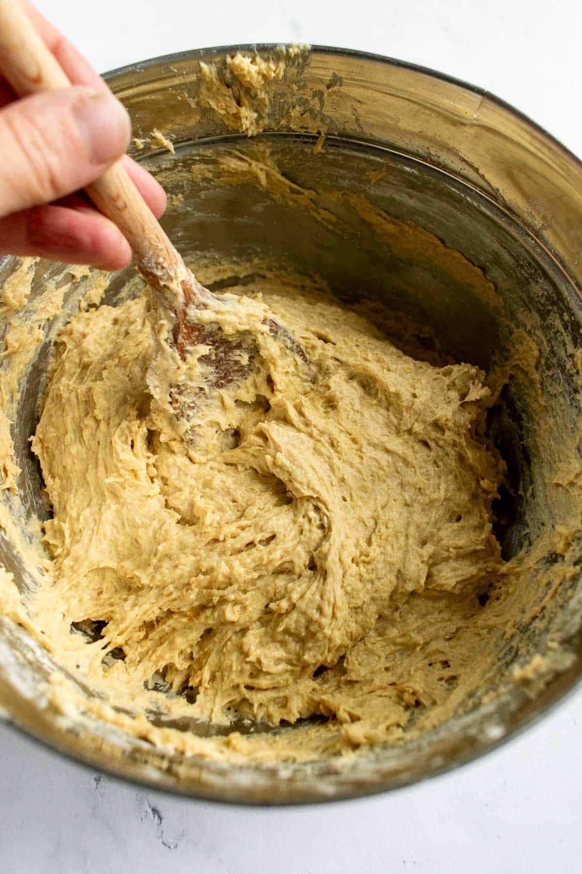 A hand mixing sticky, wet bread dough in a mixing bowl with a wooden spoon.