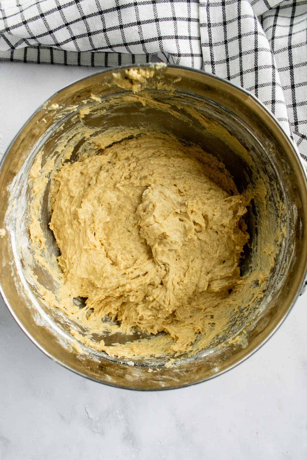 A metal bowl filled with sticky bread dough next to a black and white linen.