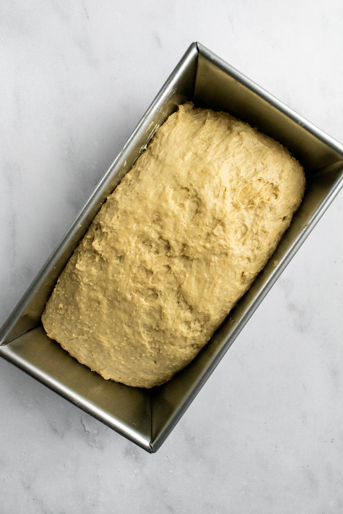 A metal loaf pan filled with a soft, beige bread dough on a light surface.