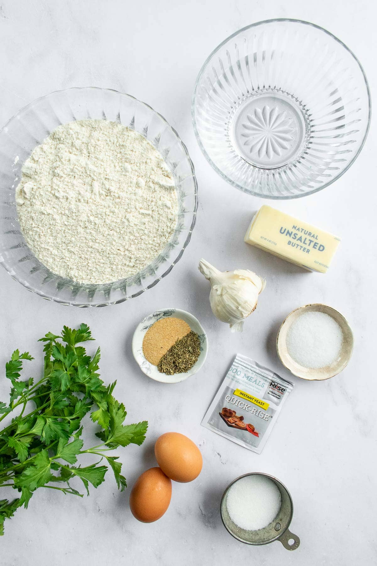 Bowls of water, flour, salt, sugar, and herbs on a table next to eggs, a head of garlic, fresh herbs, and a stick of butter.