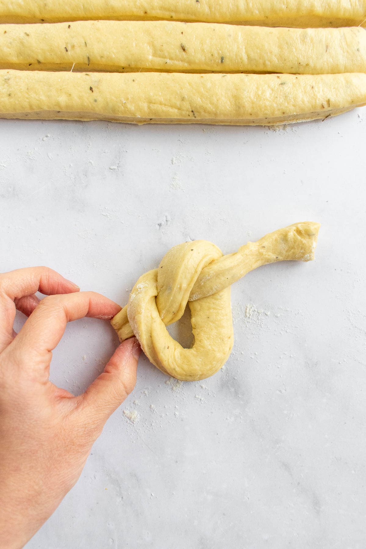 A hand tying a strand of soft bread dough into a knot on a light surface.