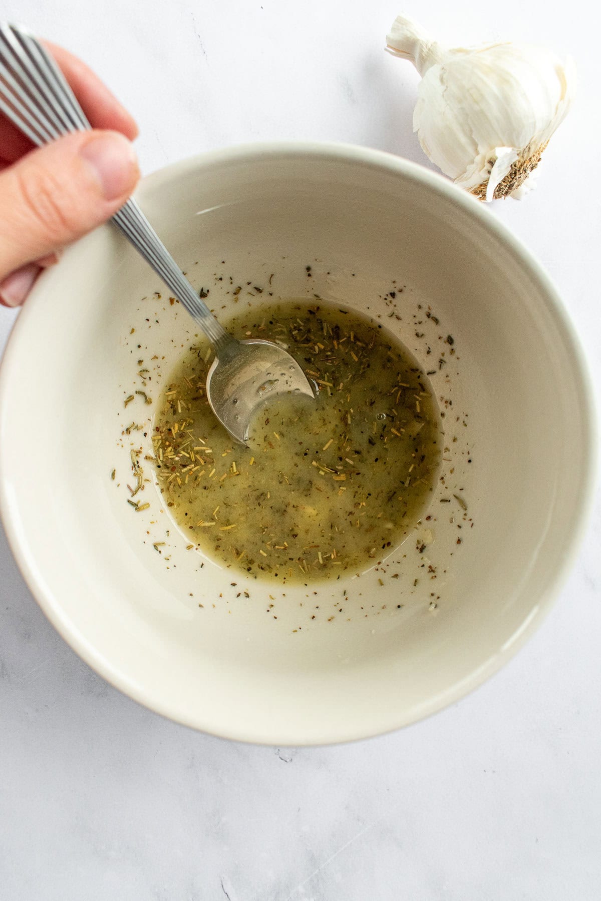 A hand mixing melted butter, dried herbs, and minced garlic in a beige bowl with a spoon.