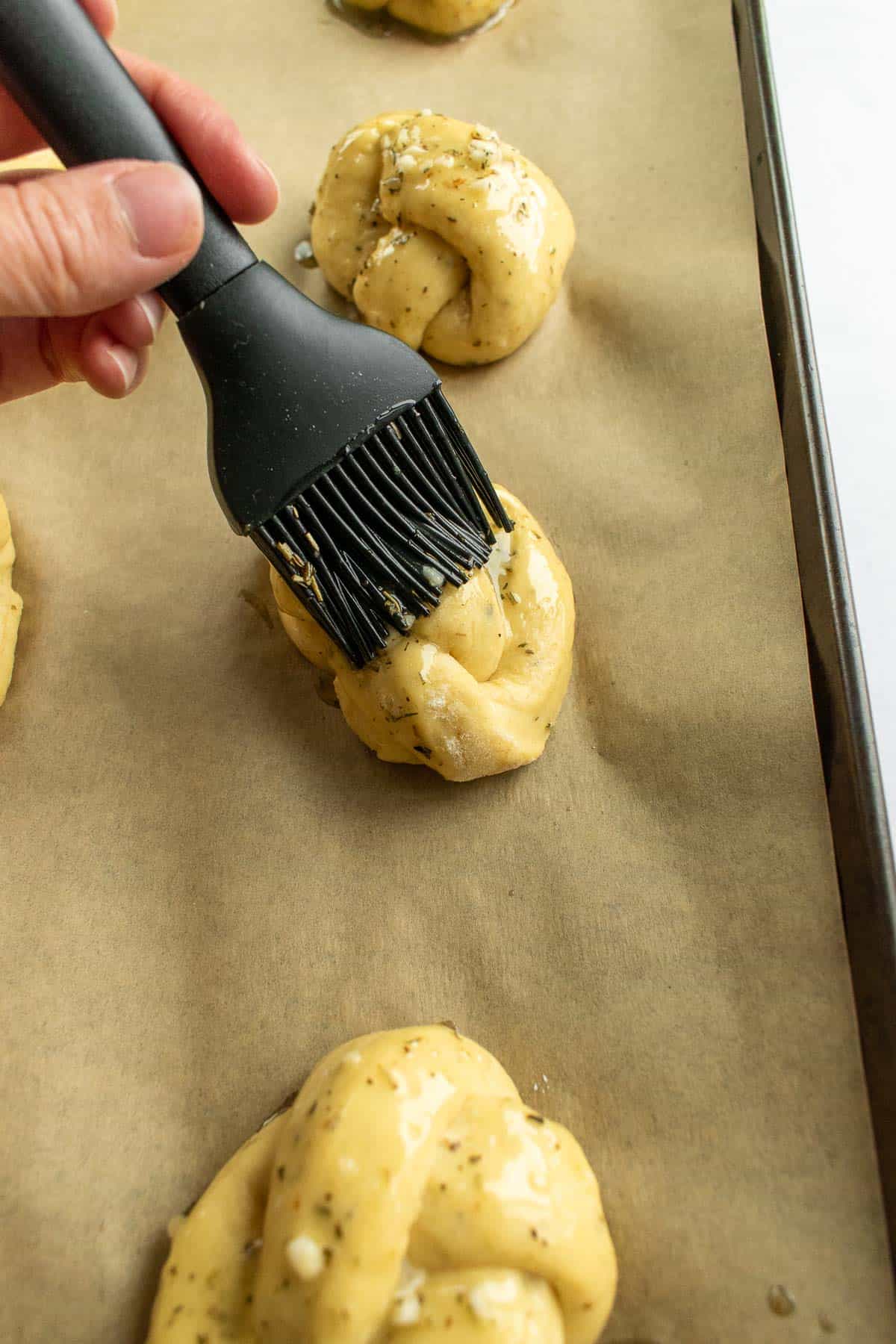 A hand brushing melted garlic butter onto unbaked garlic knots on a baking sheet lined with parchment paper.