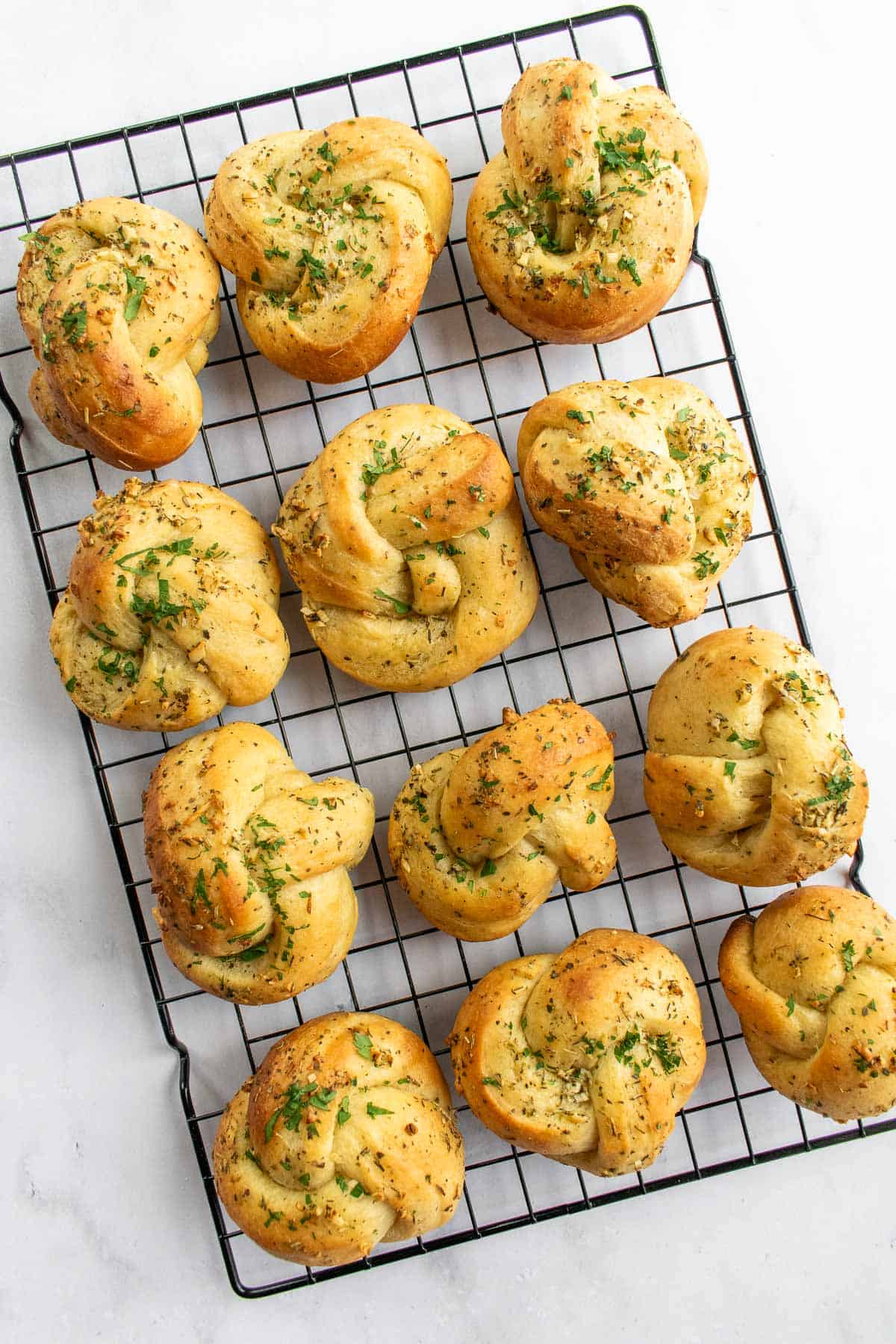 Twelve baked garlic knots on a wire cooling rack topped with melted butter and fresh chopped herbs.