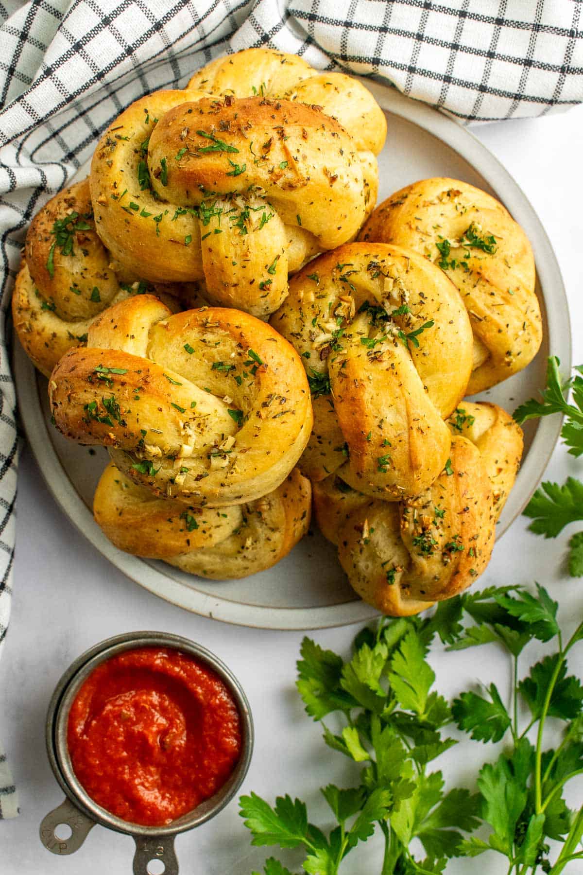 A pile of a dozen garlic knots on a grey plate next to a dish of red marinara sauce and fresh parsley.
