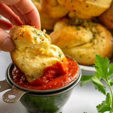 A hand dipping a garlic bread knot into a dish of red marinara sauce next to fresh parsley sprigs.