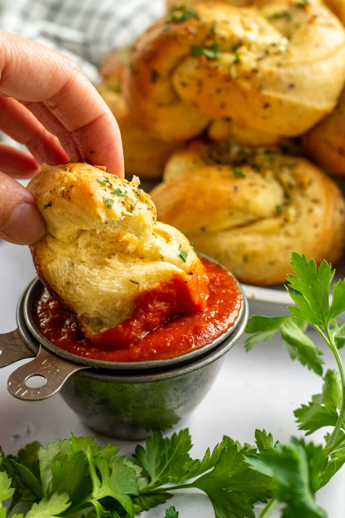 A hand dipping a garlic bread knot into a dish of red marinara sauce next to fresh parsley sprigs. 