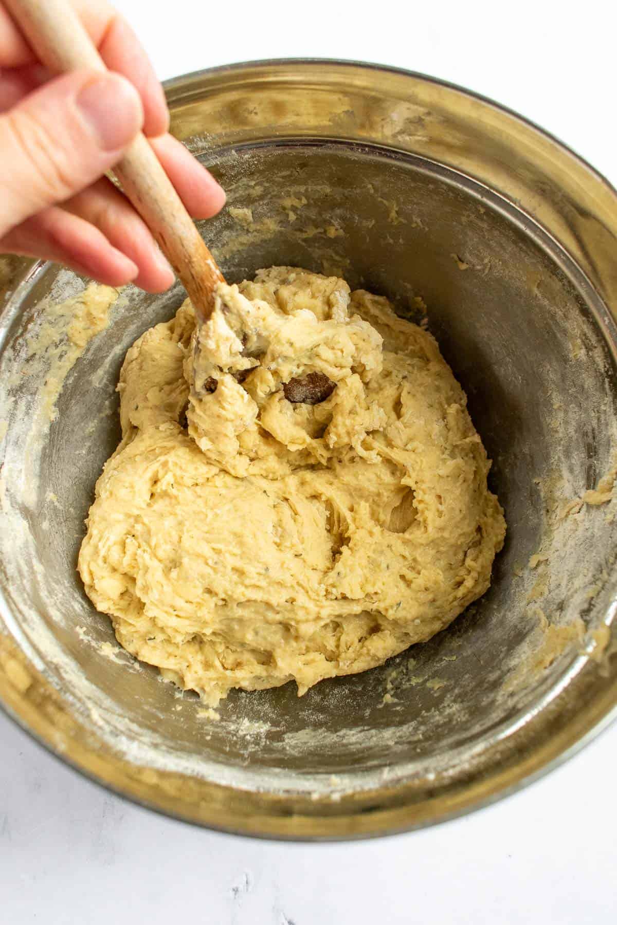A hand mixing soft bread dough in a metal bowl with a large wooden spoon.