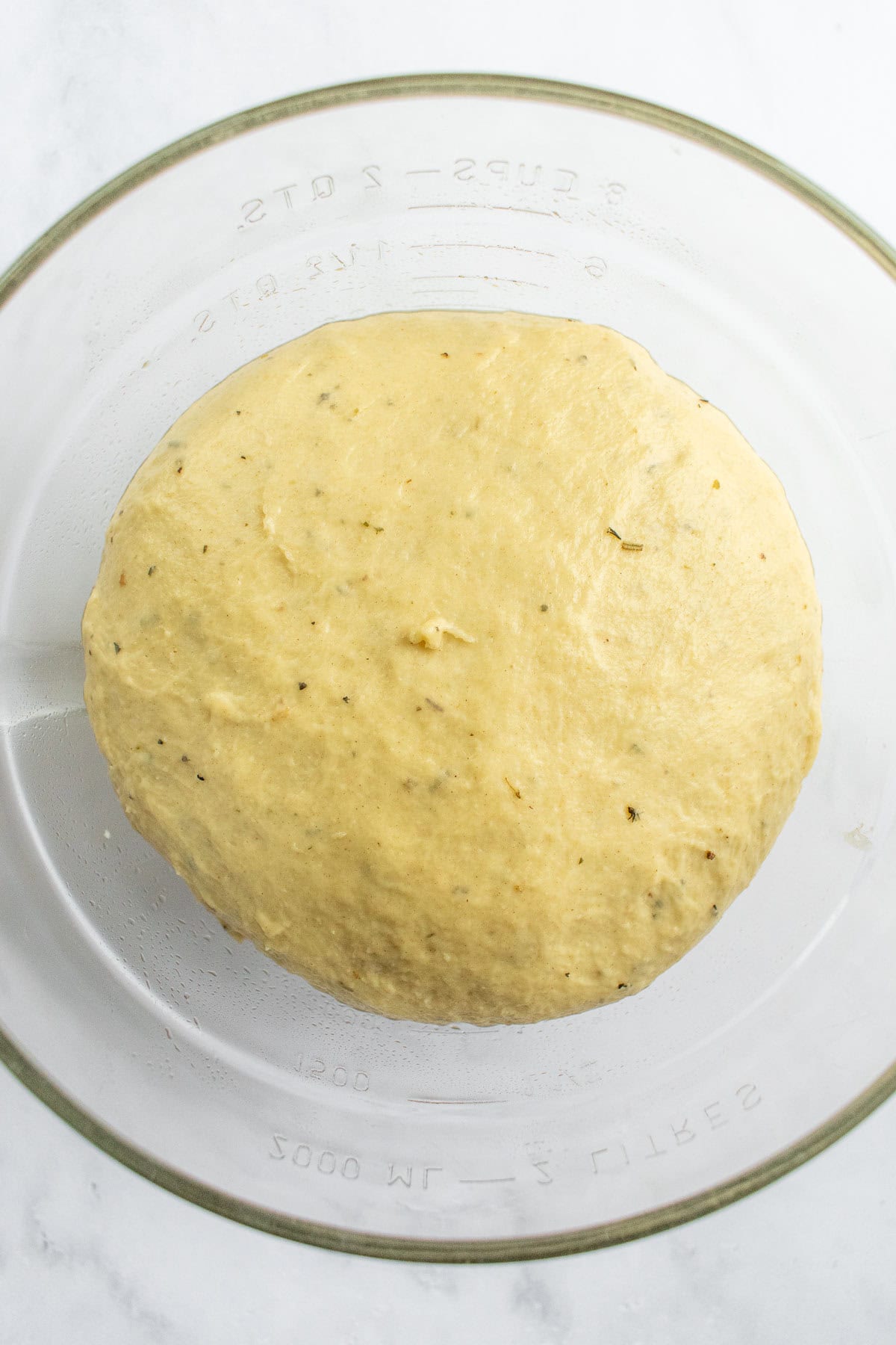 A ball of risen herb bread dough in a glass bowl.