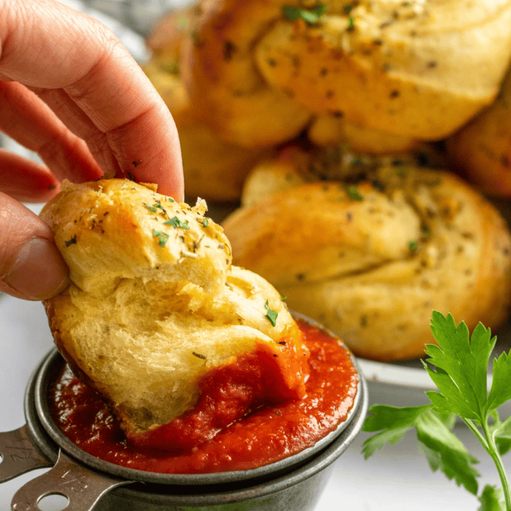 A hand dipping a torn piece of garlic bread into a metal bowl of red tomato sauce.