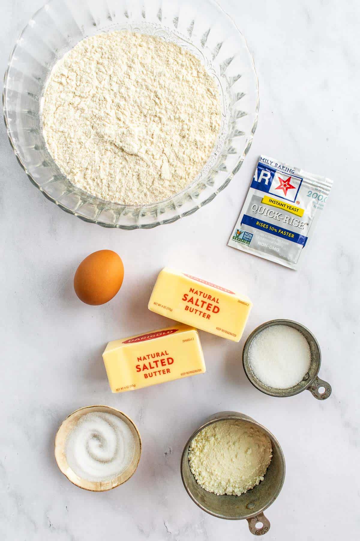 Two sticks of butter on a table next to bowls of flour, sugar, salt, and milk powder next to an egg and packet of instant yeast.