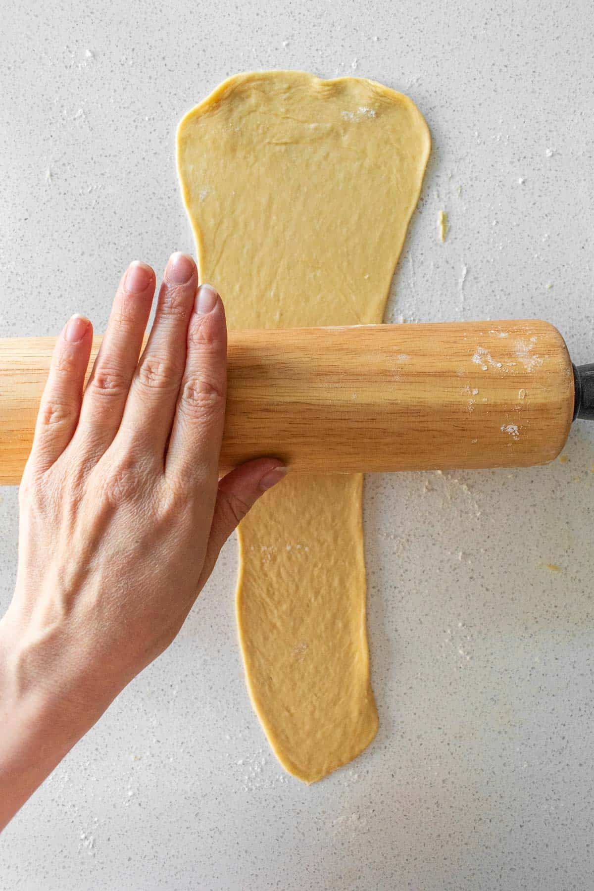 A hand rolling out a long piece of bread dough with a tapered end with a wooden rolling pin.