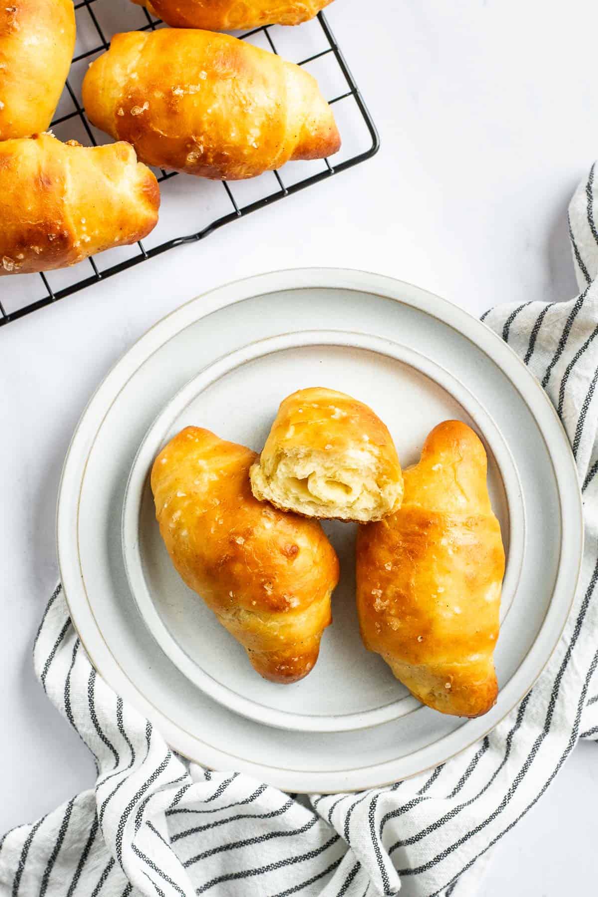 Two and a half salt bread buns on stacked grey plates next to more on a cooling rack.