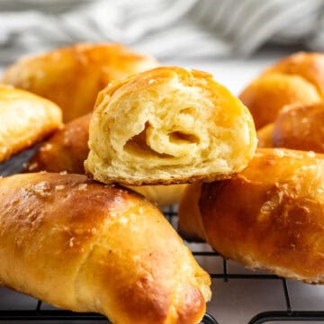 Half a dozen small bread buns on a cooling rack with one cut in half to show the fluffy texture inside.