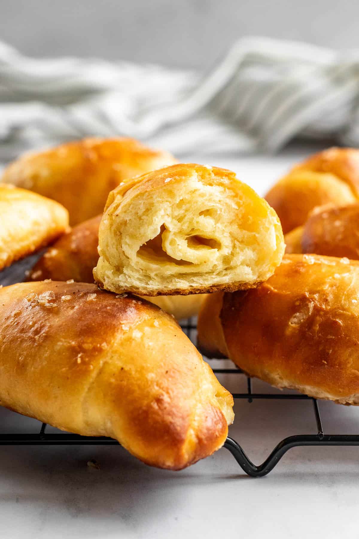 Half a dozen salt bread bread buns on a cooling rack with one cut in half to show the fluffy texture inside.