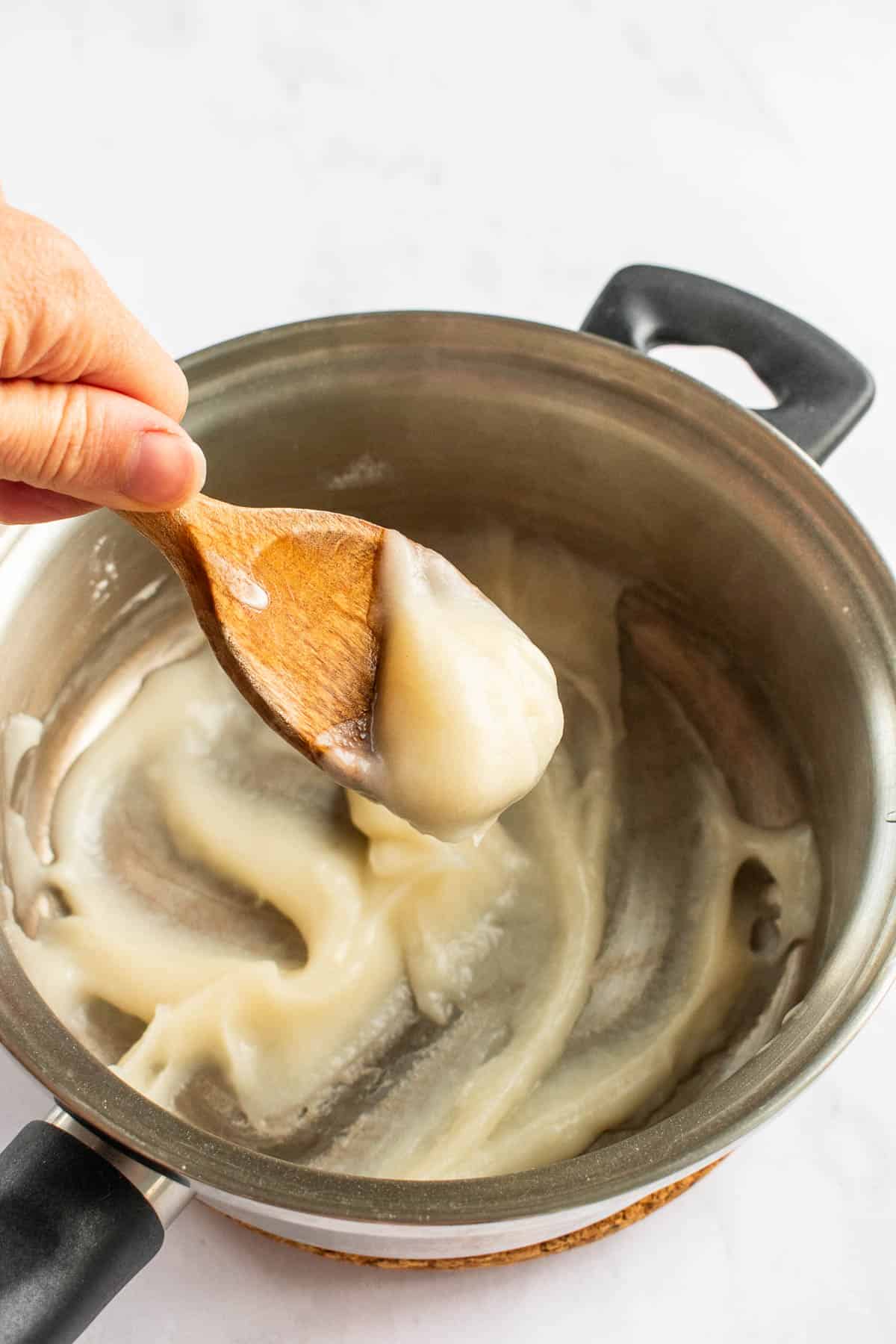 A hand mixing a thick roux of flour and water in a saucepan with a wooden spoon.