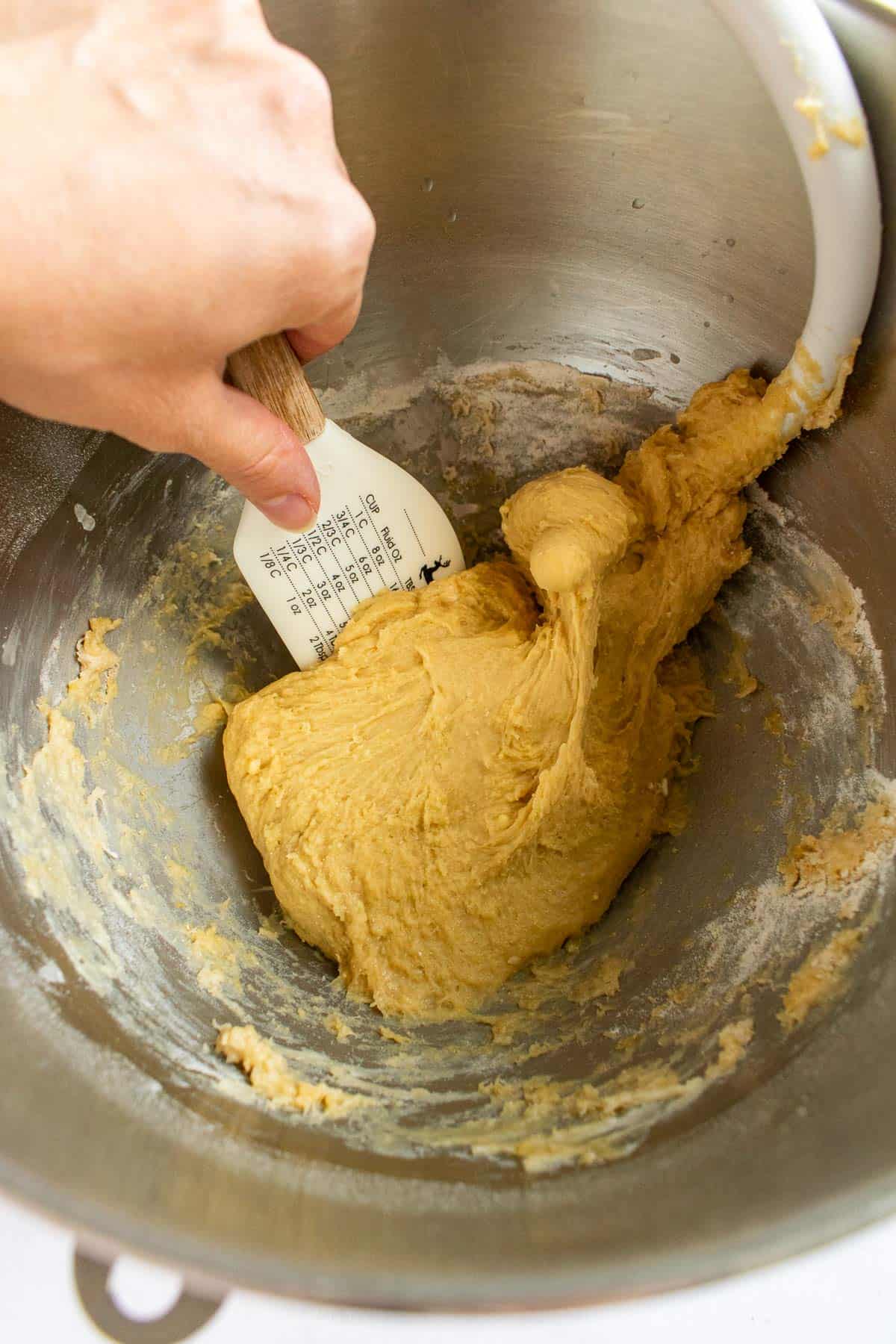 A hand scooping bread dough off the side of a mixing bowl with a silicone spatula.