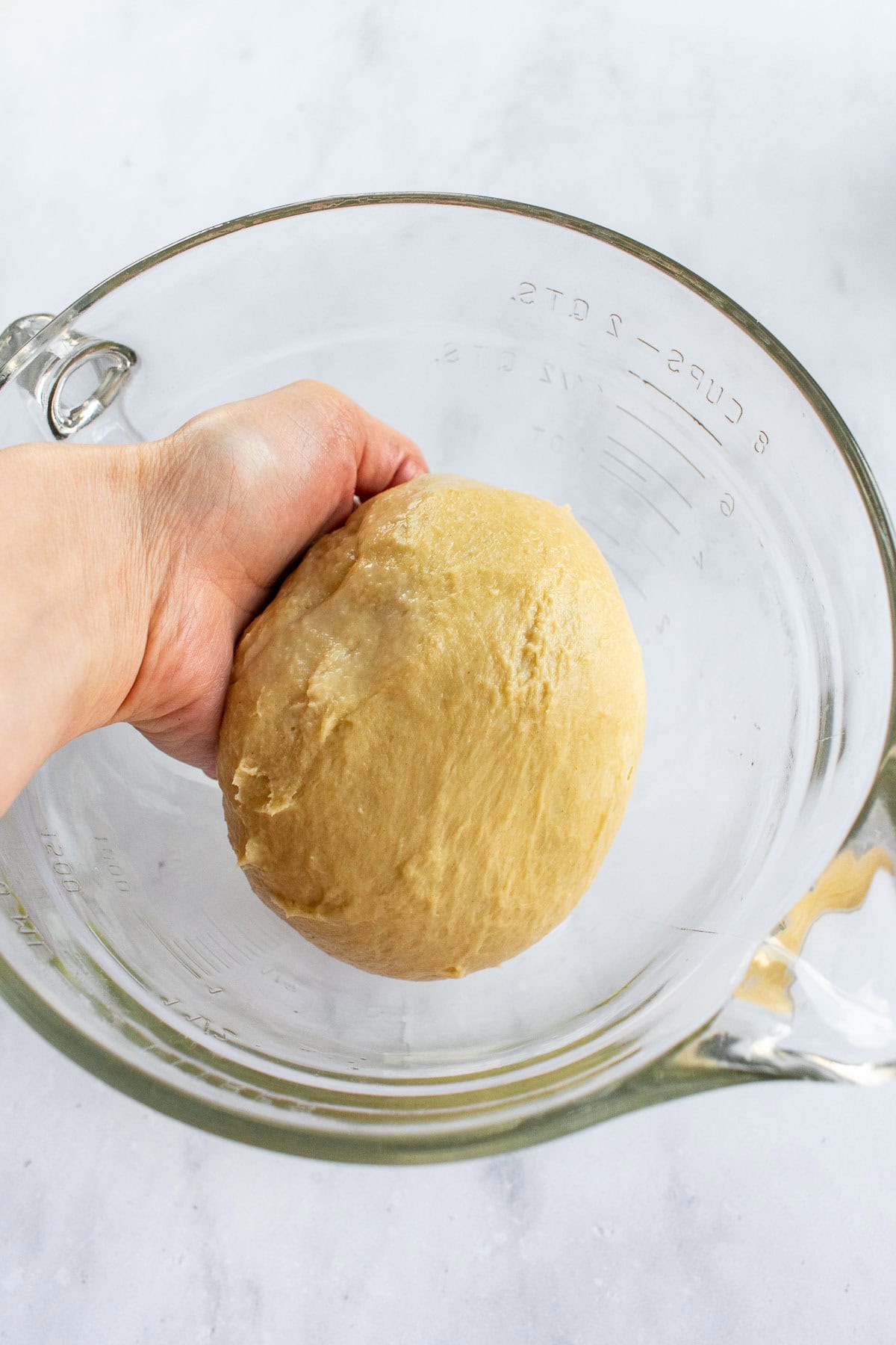 A hand placing a ball of sticky bread dough in a glass mixing bowl.