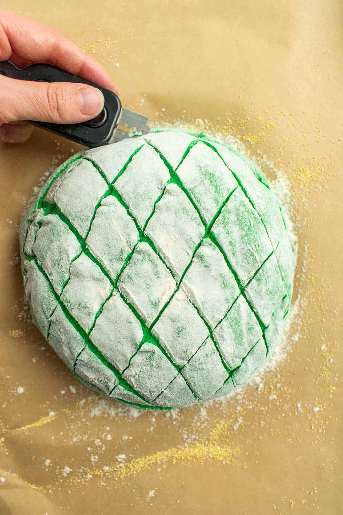 A hand scoring a ball of green bread dough with a cross-hatch pattern.