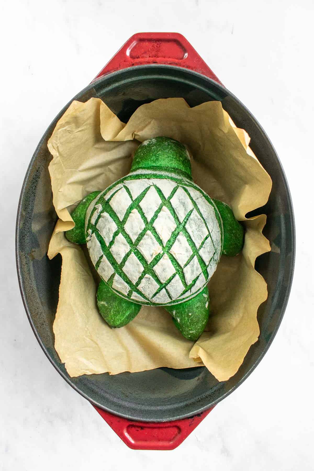 A green loaf of turtle shaped bread inside a red Dutch oven on parchment paper.