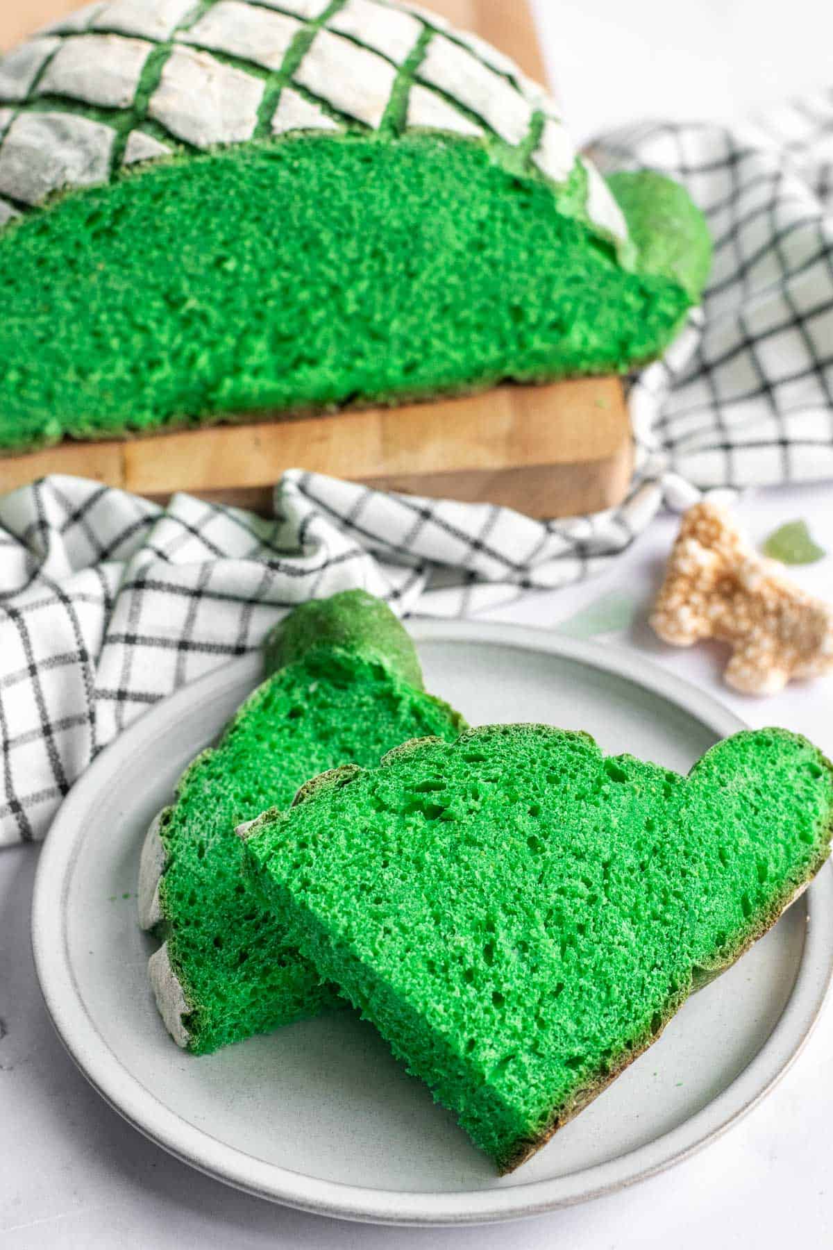 Two slices of green bread on a grey plate next to the rest of a green loaf on a wood board.