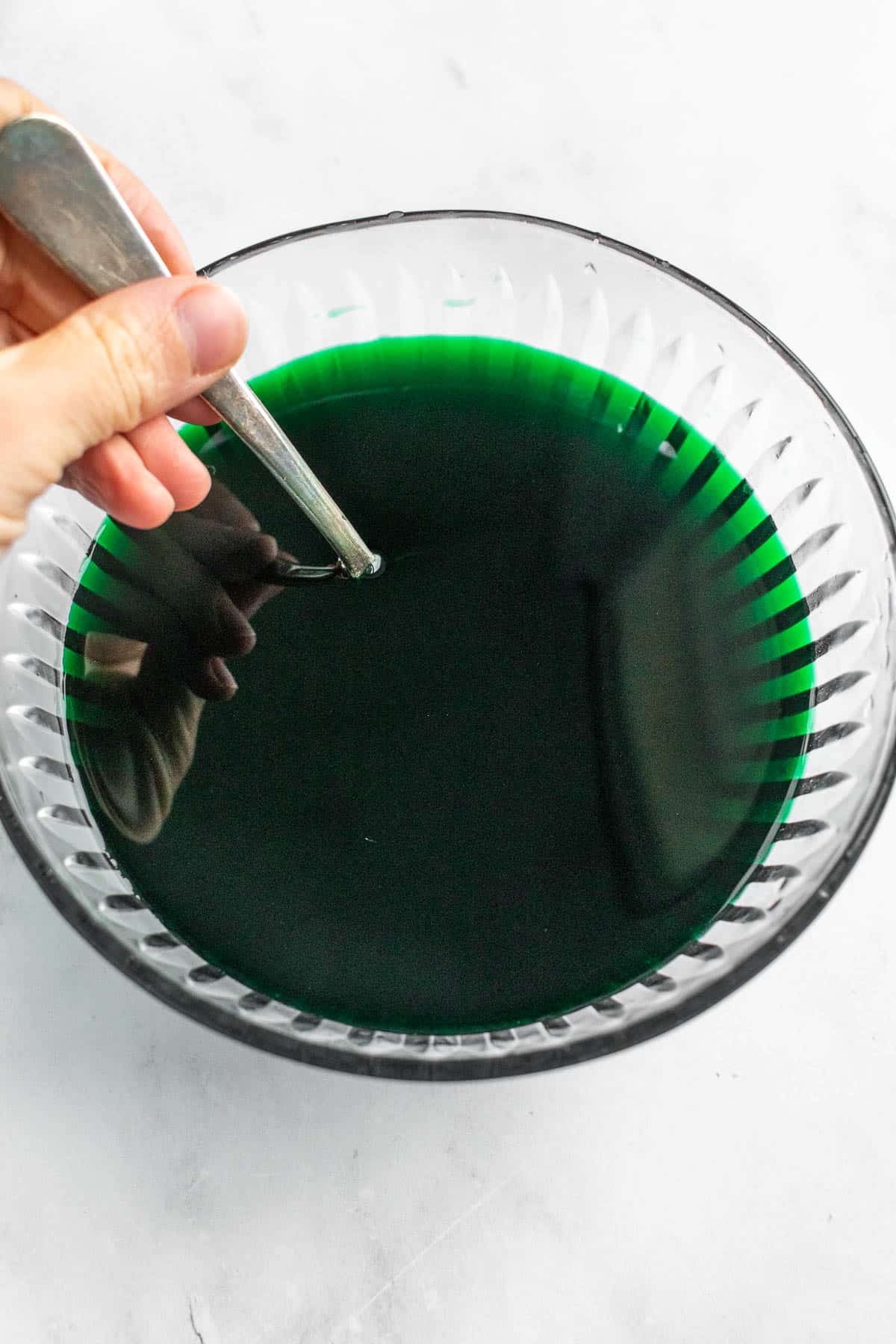 A hand stirring dark green water in a glass bowl with a metal spoon.