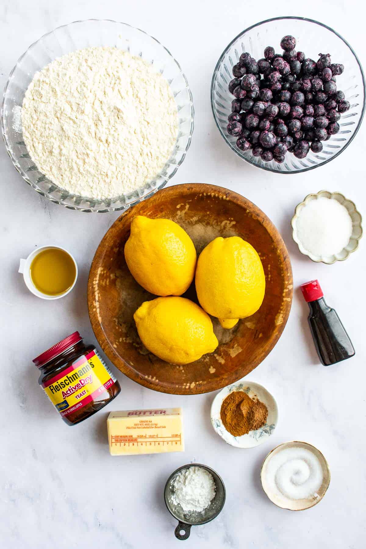 Three lemons in a bowl next to bowls of flour, frozen blueberries, salt, sugar, olive oil, and cinnamon on a light table.