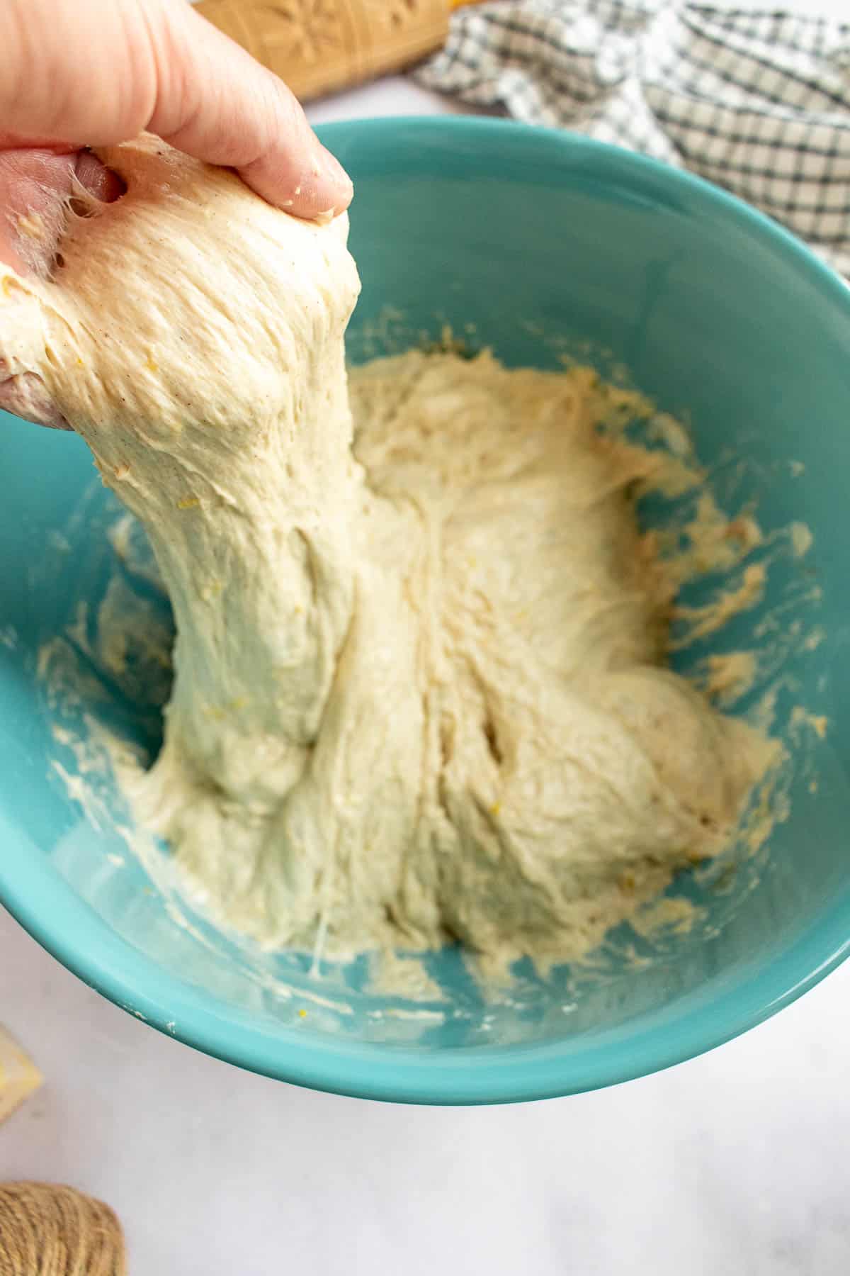 A hand pulling up a handful of sticky bread dough from a mixing bowl.