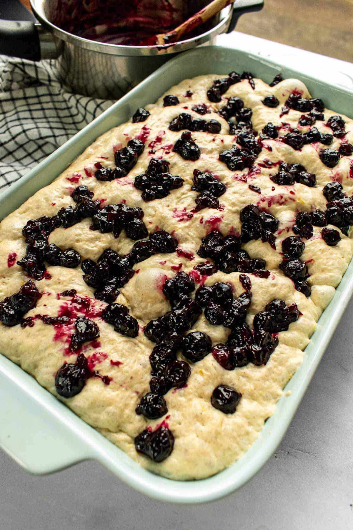 Cooked blueberries sitting on bubbly focaccia dough in a baking dish on a table.
