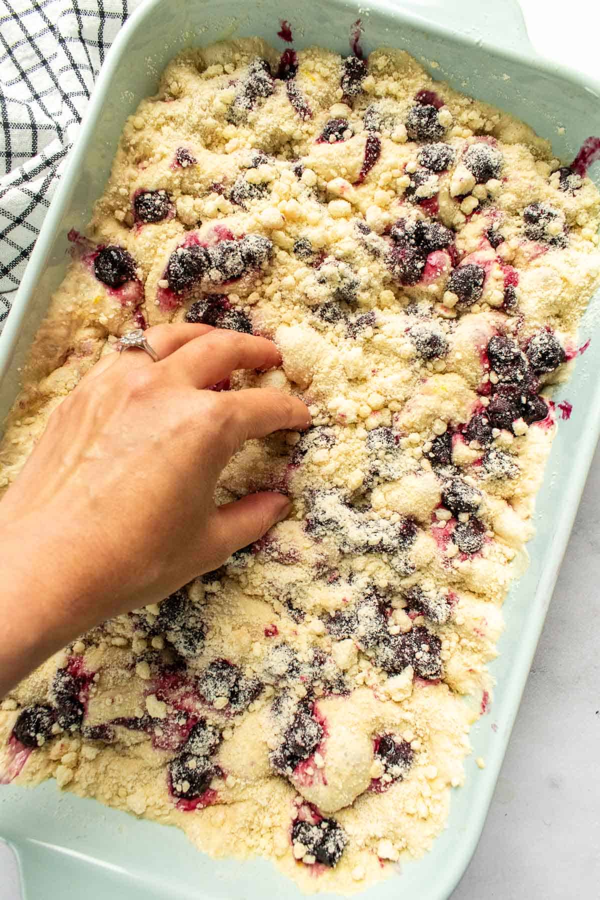 A hand pressing dimples into blueberry lemon focaccia dough in a light blue baking dish.