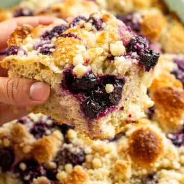 A hand holding a large piece of focaccia studded with blueberries and topped with crumbly streusel.