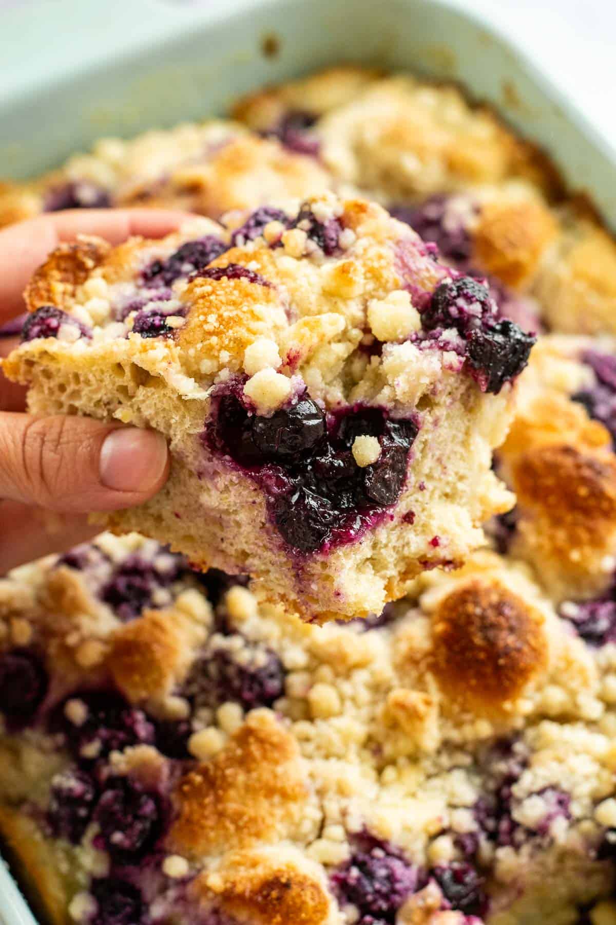 A hand holding a large piece of focaccia studded with blueberries and topped with crumbly streusel. 