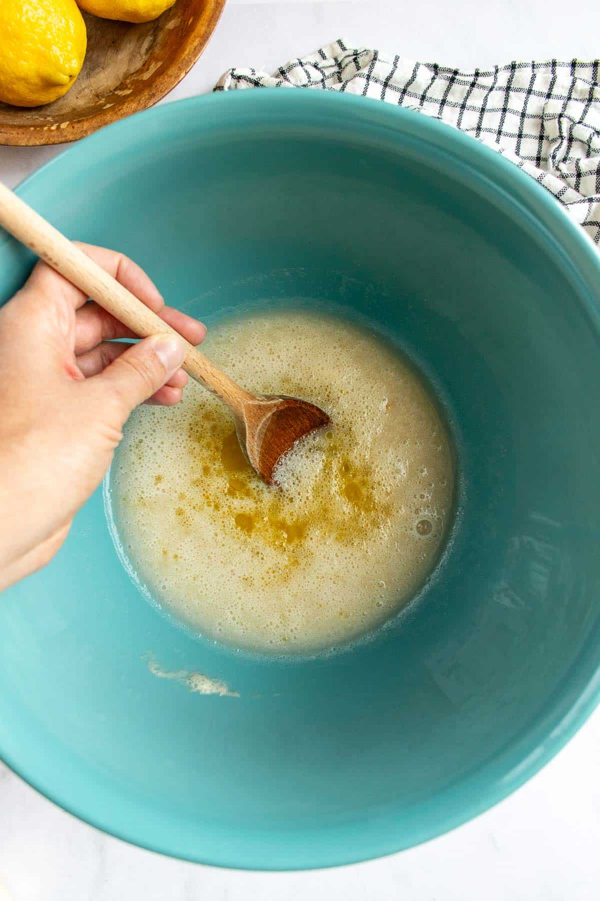 A hand stirring olive oil into water and yeast with a large wooden spoon.