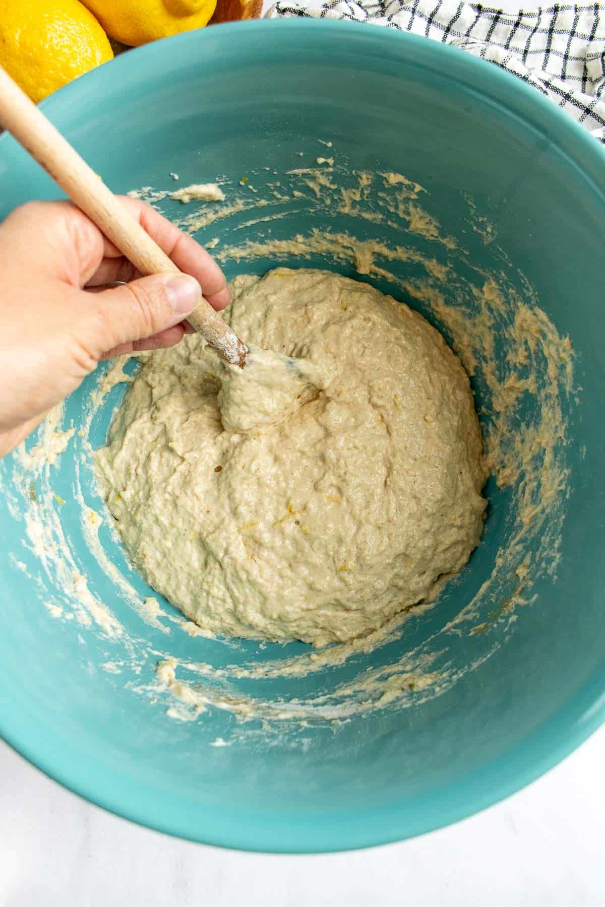A hand stirring wet bread dough in a blue bowl with a large wooden spoon.
