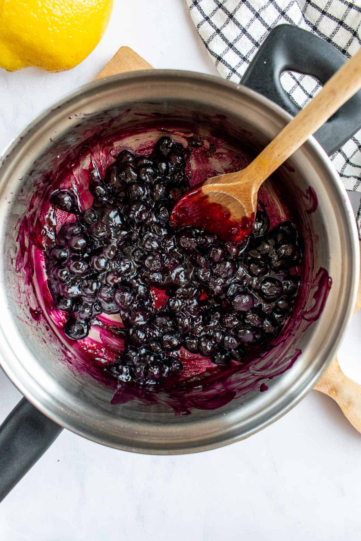 A thick blueberry filling in a metal saucepan next to a lemon on a table.