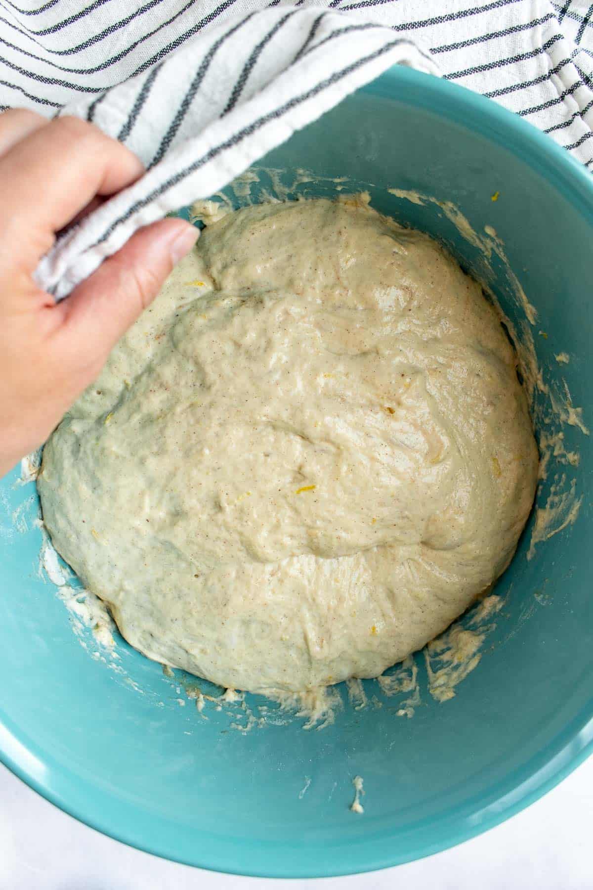 A hand holding a black and white linen over a mixing bowl filled with wet, sticky bread dough.