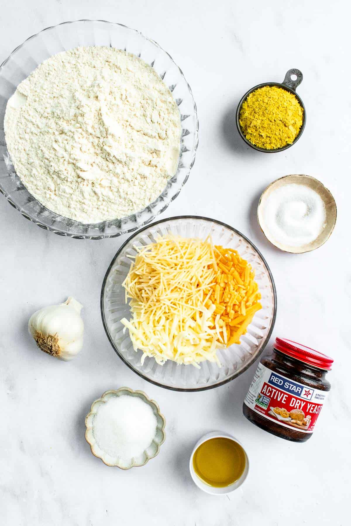 Bowls of flour, salt, shredded cheese, sugar, olive oil, and nutritional yeast next to a jar of yeast and a head of garlic.