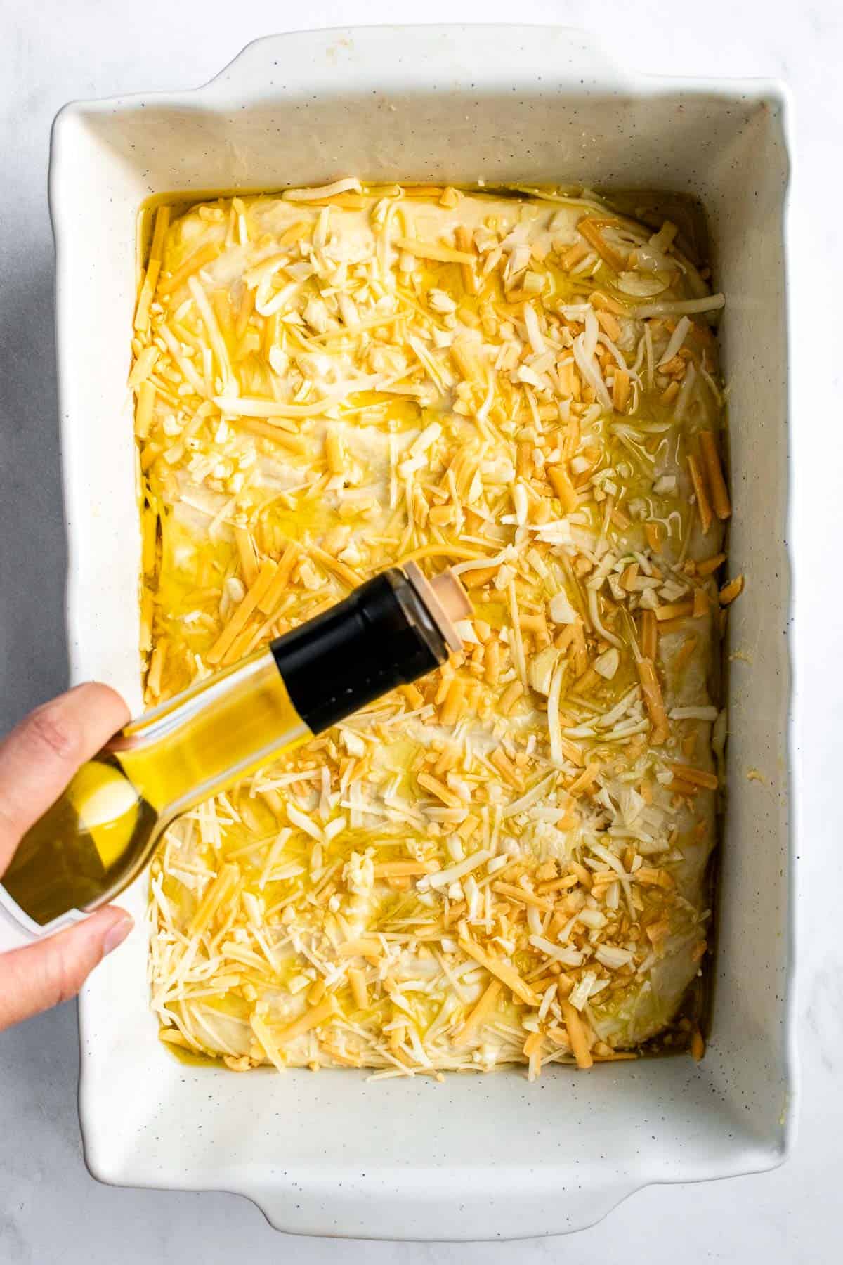 A hand drizzling olive oil from a bottle onto focaccia dough sprinkled with shredded cheese.