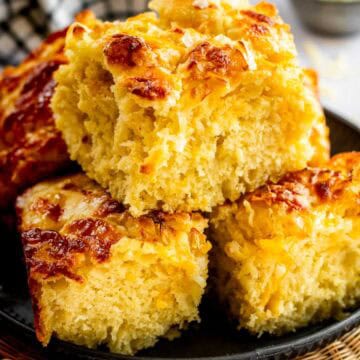 Three large square pieces of fluffy cheese focaccia piled on a metal plate.