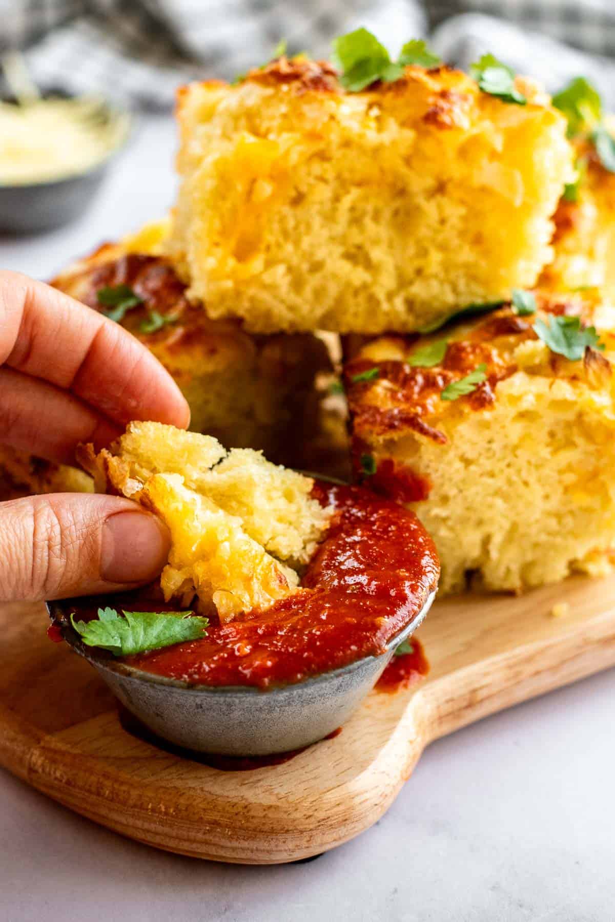 A hand dunking a torn piece of focaccia bread into a metal cup of red marinara sauce. 