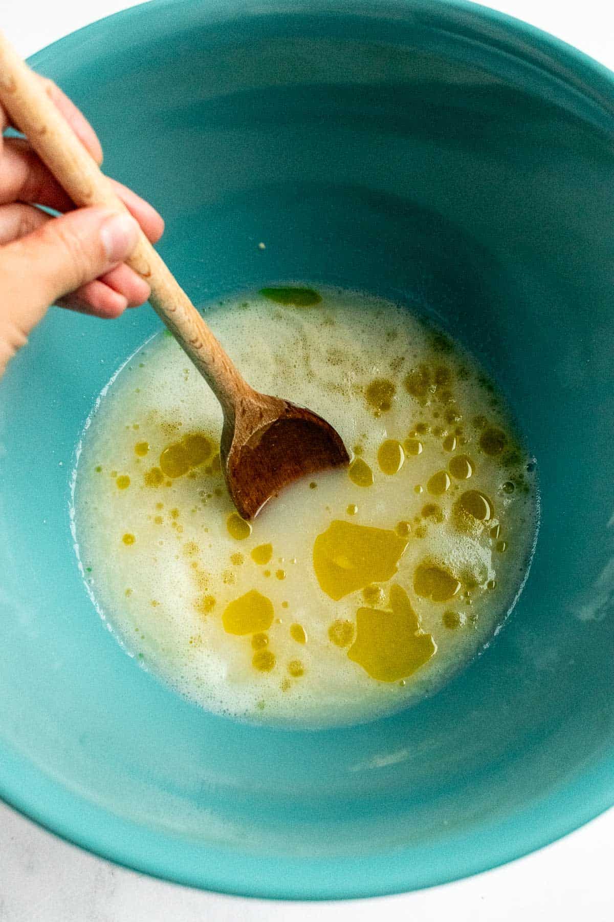 A hand stirring frothy yeast and olive oil in water in a bowl with a wooden spoon.