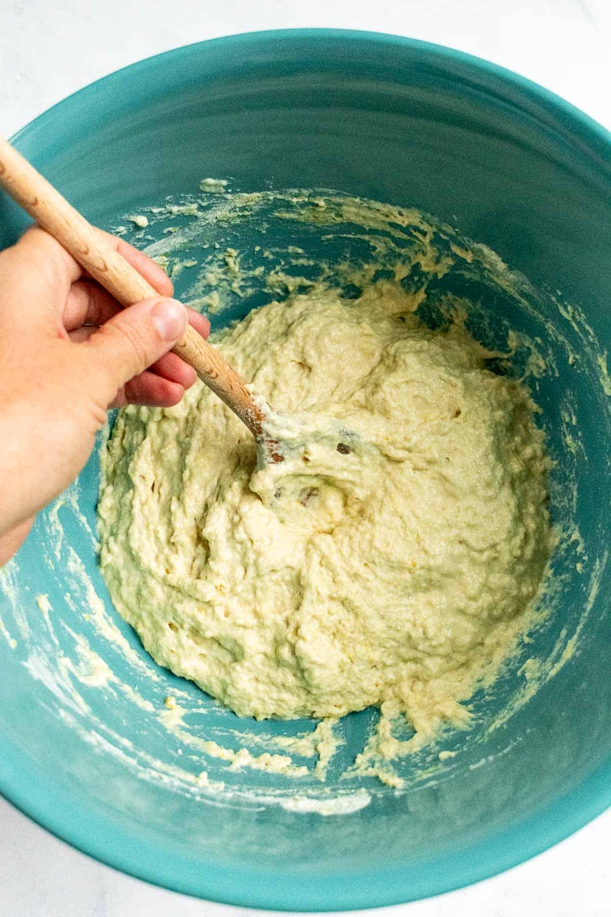 A hand stirring wet bread dough in a blue mixing bowl with a wooden spoon.