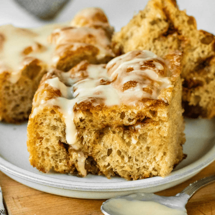 Three large squares of cinnamon roll focaccia on a plate with a cinnamon swirl filling and glaze drizzled on top.