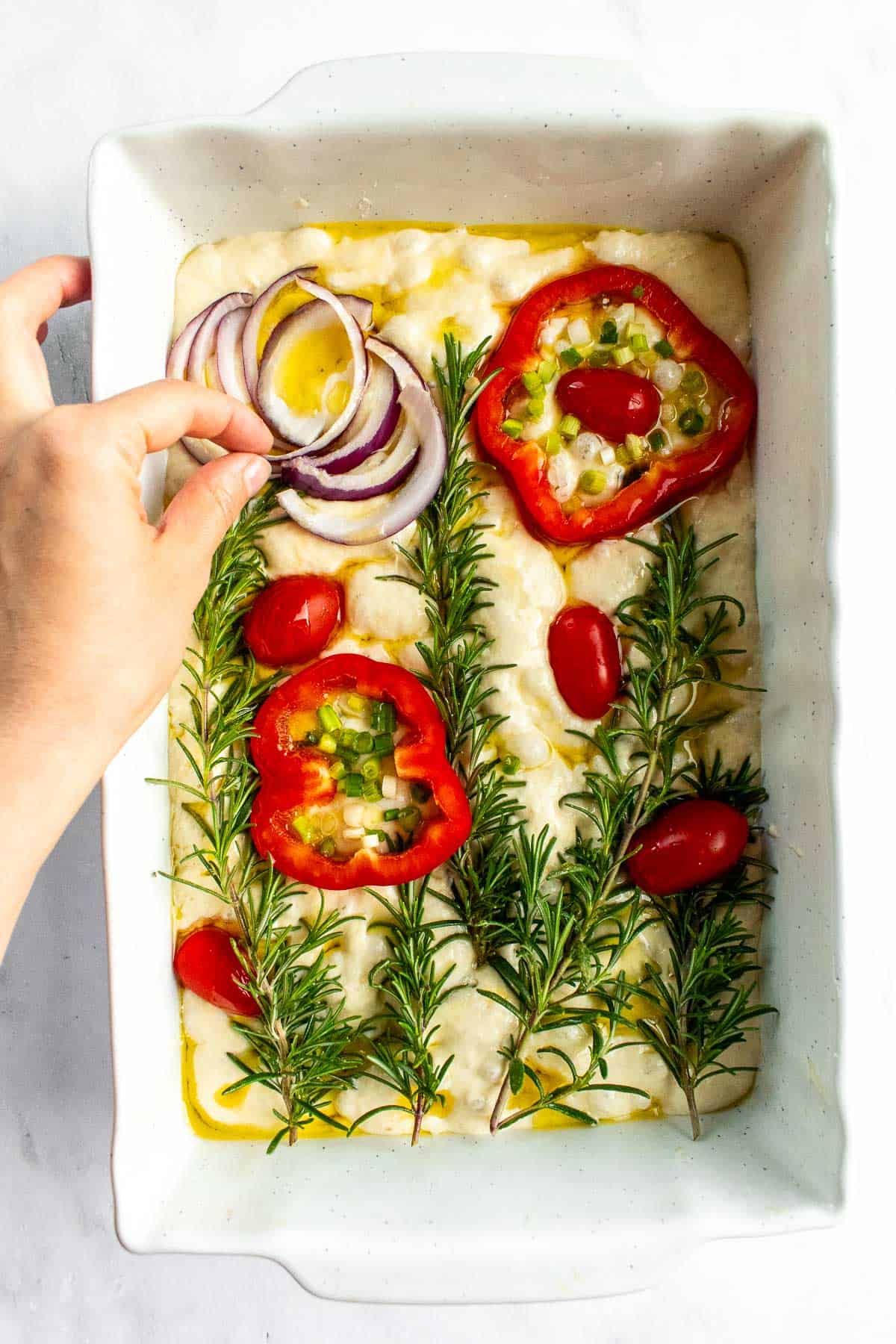 Focaccia dough in a white baking dish being decorated with raw red onion, red bell pepper, cherry tomatoes, and rosemary.