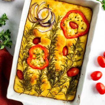 A white baking dish with focaccia bread topped with roasted vegetables and herbs.