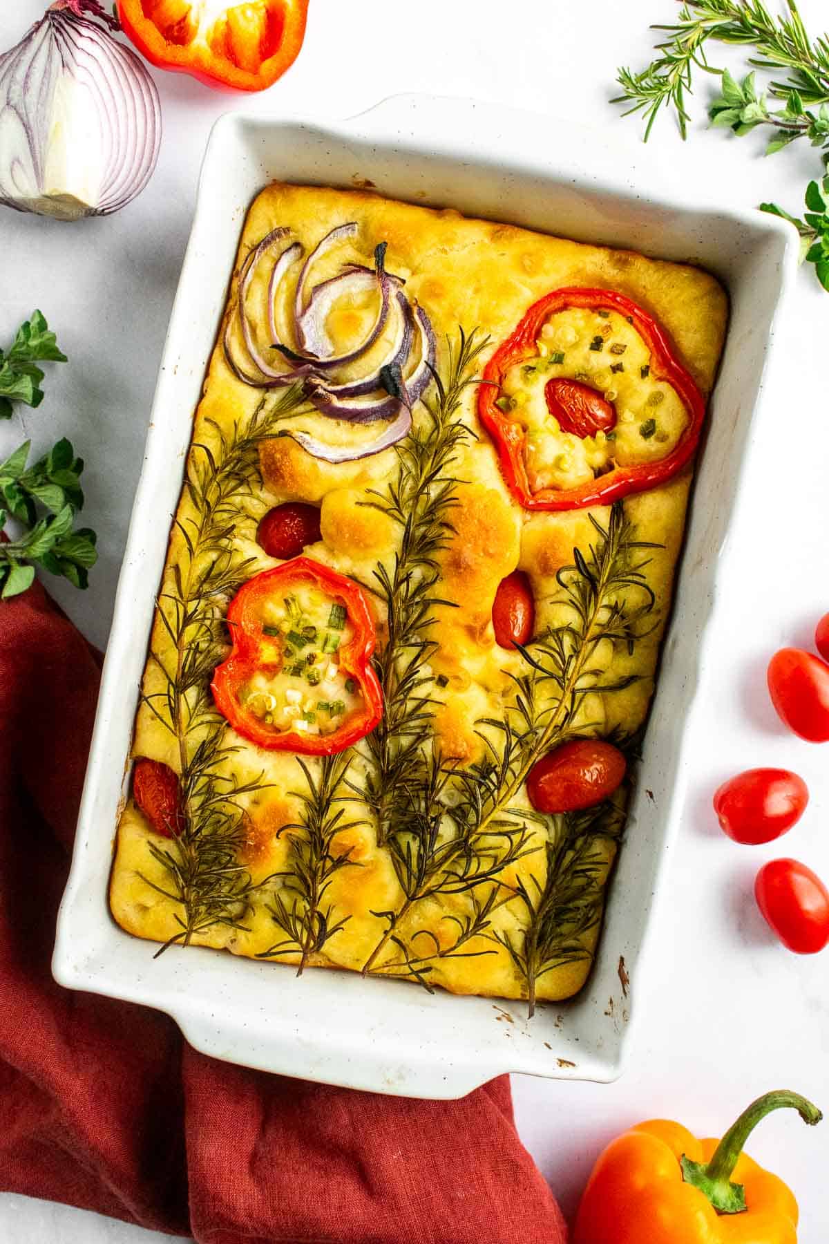 A white baking dish with focaccia bread topped with roasted vegetables and herbs.